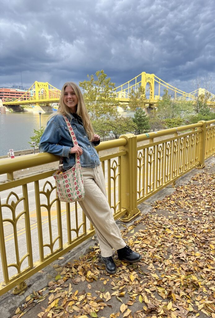 Ashley leaning against yellow railing, wearing a stylish outfit.