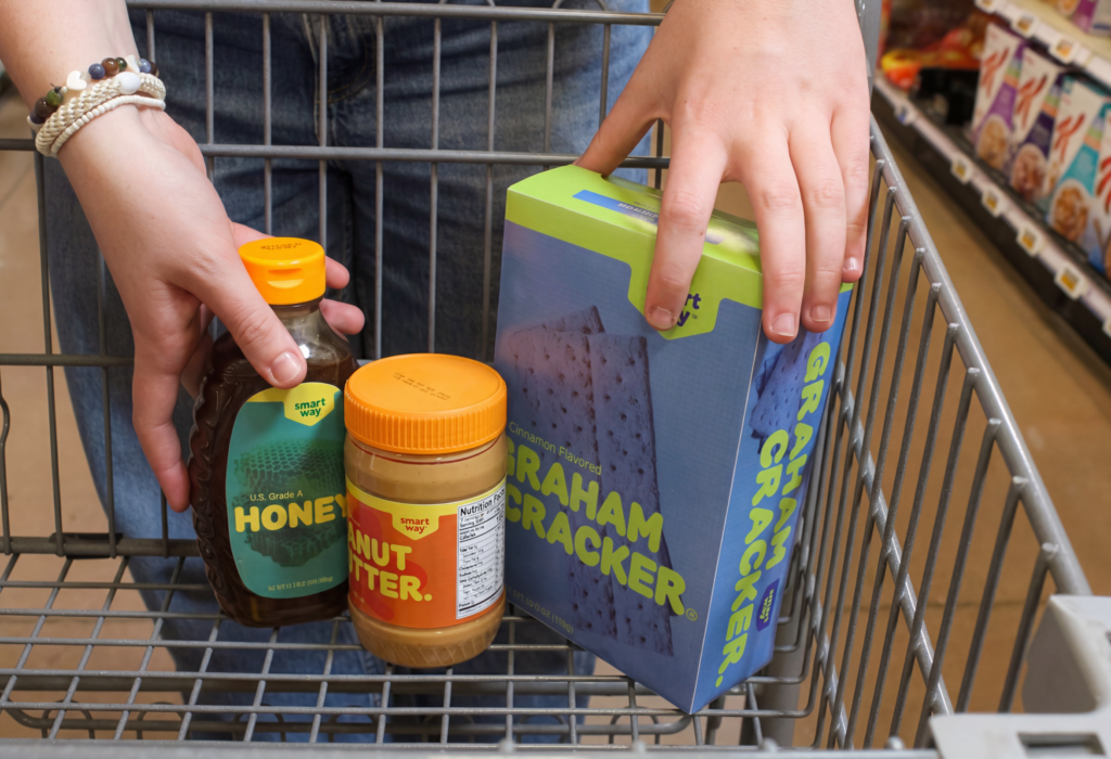 Grocery cart filled with colorful Smart Way pantry products in a bright grocery store setting.